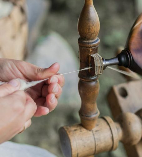 Hands working a wooden spinning wheel, guiding thread through a guide.