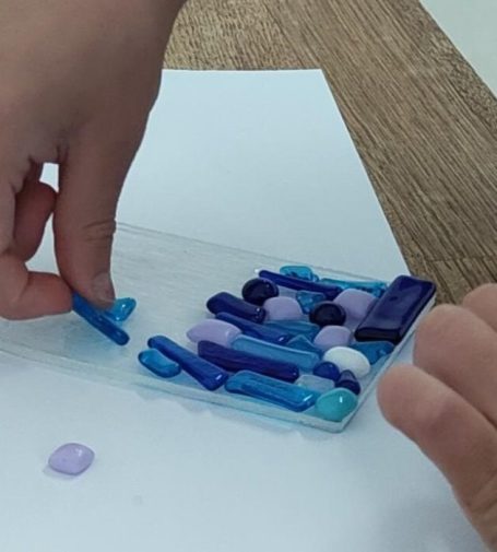 Hands arranging various coloured bits of glass on a clear glass surface.
