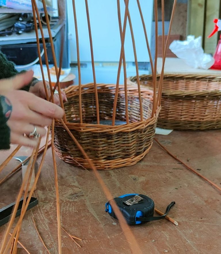 Hands weaving thin strips of willow into a circular basket on a wooden table.